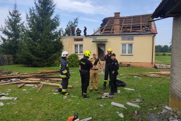 Lubelski Komendant Wojewódzki rozmawiający ze strażakami. Za nimi pozbawiony dachu dom jednorodzinny. Wokół porozrzucane elementy dachu.