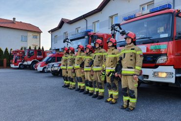 Strażacy podczas uroczystej zbiórki na tle pojazdów pożarniczych